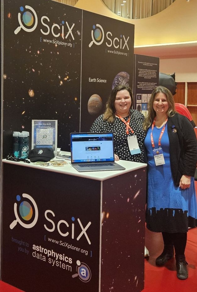 Photo of a SciX outreach booth at a conference. Two women wearing conference badges stand smiling behind a exhibit counter decorated with the SciX logo and the phrase "brought to you by astrophysics data system." Behind them are tall black display panels featuring space imagery and the SciX logo and URL. On the counter is an open laptop showing the SciX Science Explorer homepage, along with promotional items such as water bottles, a hat, buttons, and a flyer. 