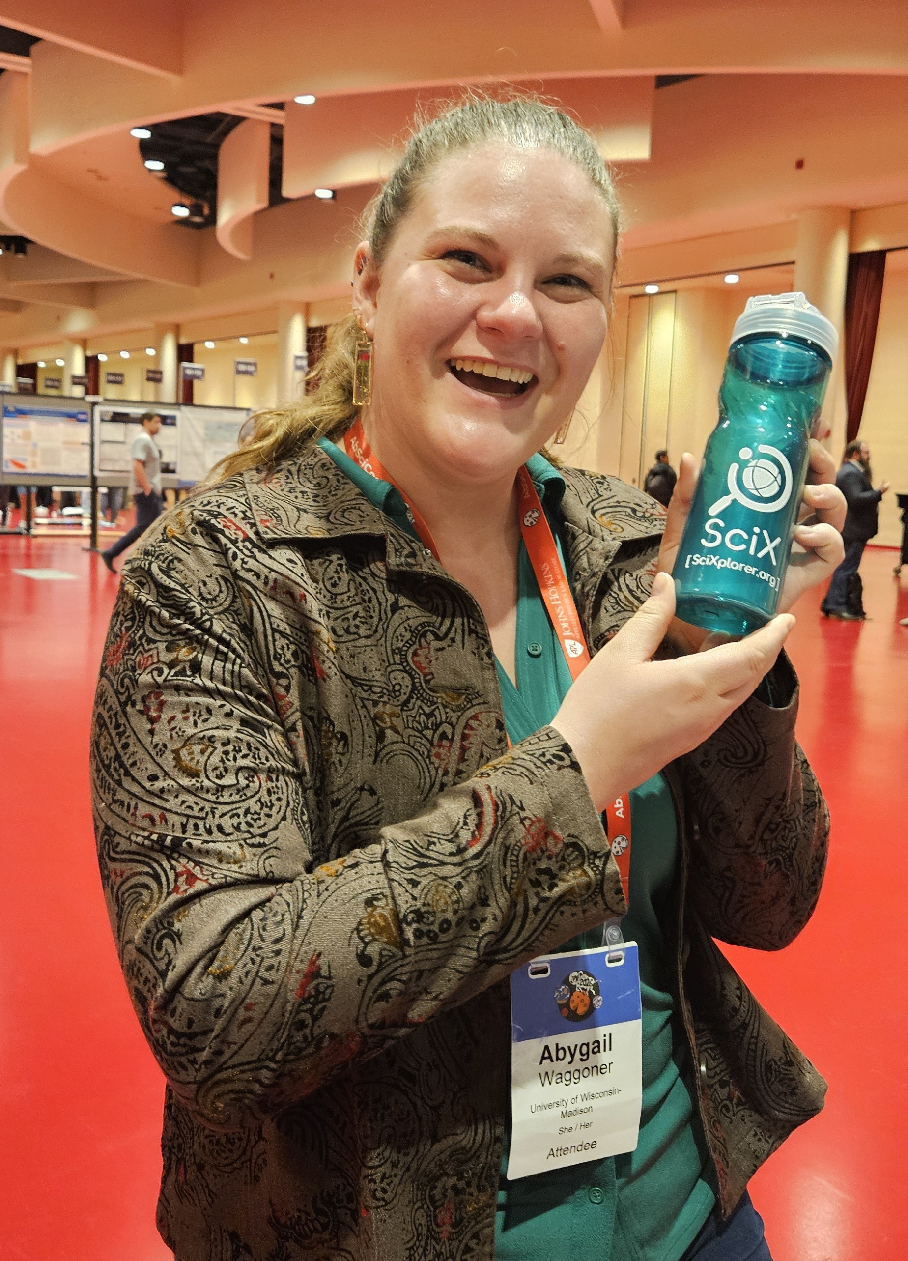 Smiling woman in a patterned jacket and orange conference lanyard stands in a bright exhibit hall with red floors, holding up a teal water bottle printed with the SciX logo and "SciXplorer.org."