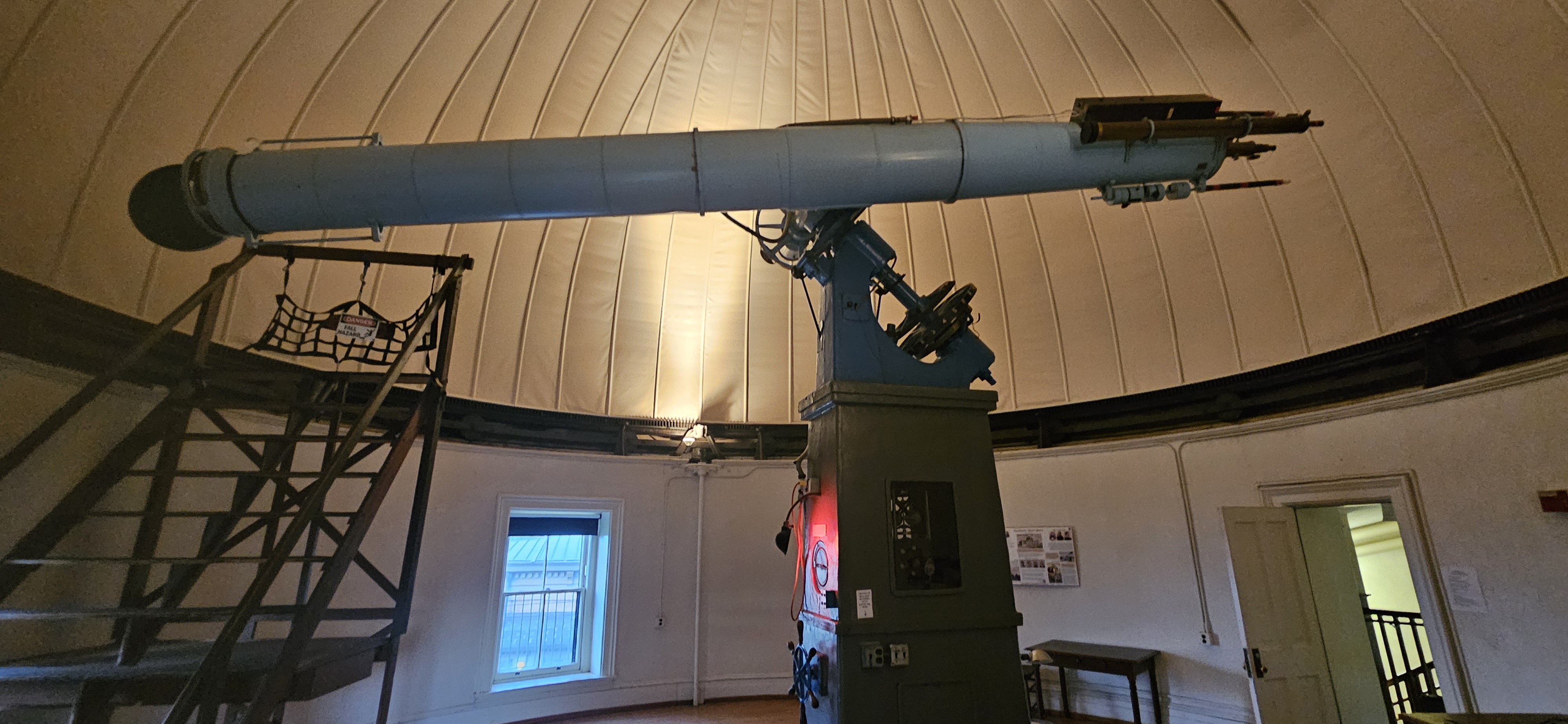 Large, vintage refracting telescope mounted on a tall pedestal inside a domed observatory room. A wooden moveable way with a small observing platform and safety net stands to the left of the long pale-blue telescope tube, which extends nearly wall to wall beneath the curved, ribbed dome ceiling. A window, doors, and a small table line the circular wall below the dome.