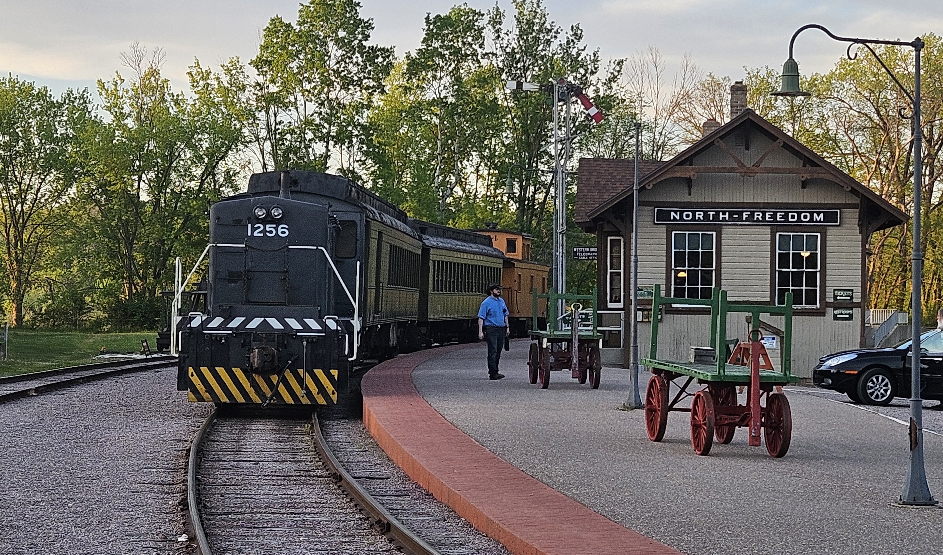 A black diesel locomotive numbered 1256 is stopped on curved tracks beside a small historic train station labeled "North Freedom," with green baggage carts on the platform, a few people walking nearby, a parked car to the side, and leafy trees in the background under a soft evening sky.