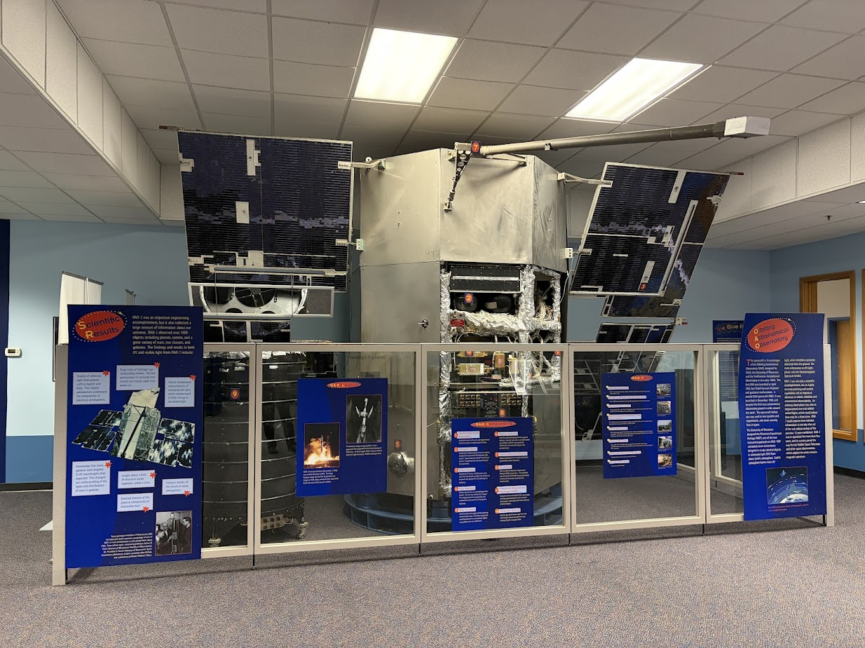 Full-scale spacecraft model displayed indoors behind a low glass barrier, with large dark solar panels extended on either side and instrument components visible in the central body. Bright blue informational panels with text, photos, and diagrams are arranged along the barrier, explaining the satellite's scientific results and mission details. The exhibit sits in a carpeted room with fluorescent ceiling lights and light blue walls.