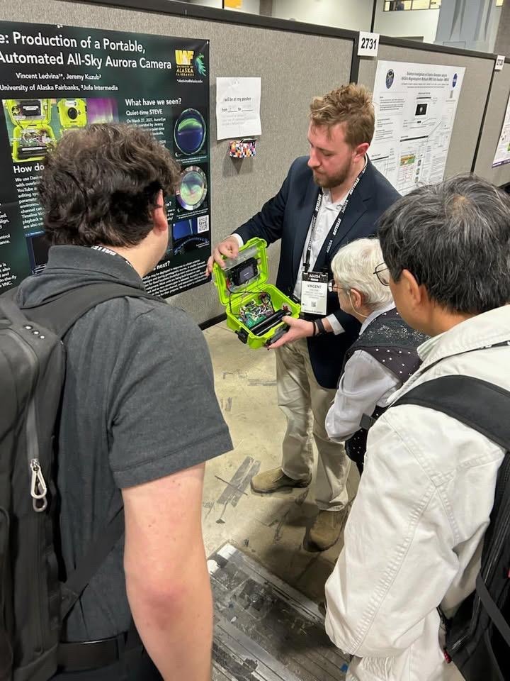 Conference attendees gather around a poster presentation while presenter Vincent Ledvina demonstrates a portable all-sky aurora camera housed in a bright green case.