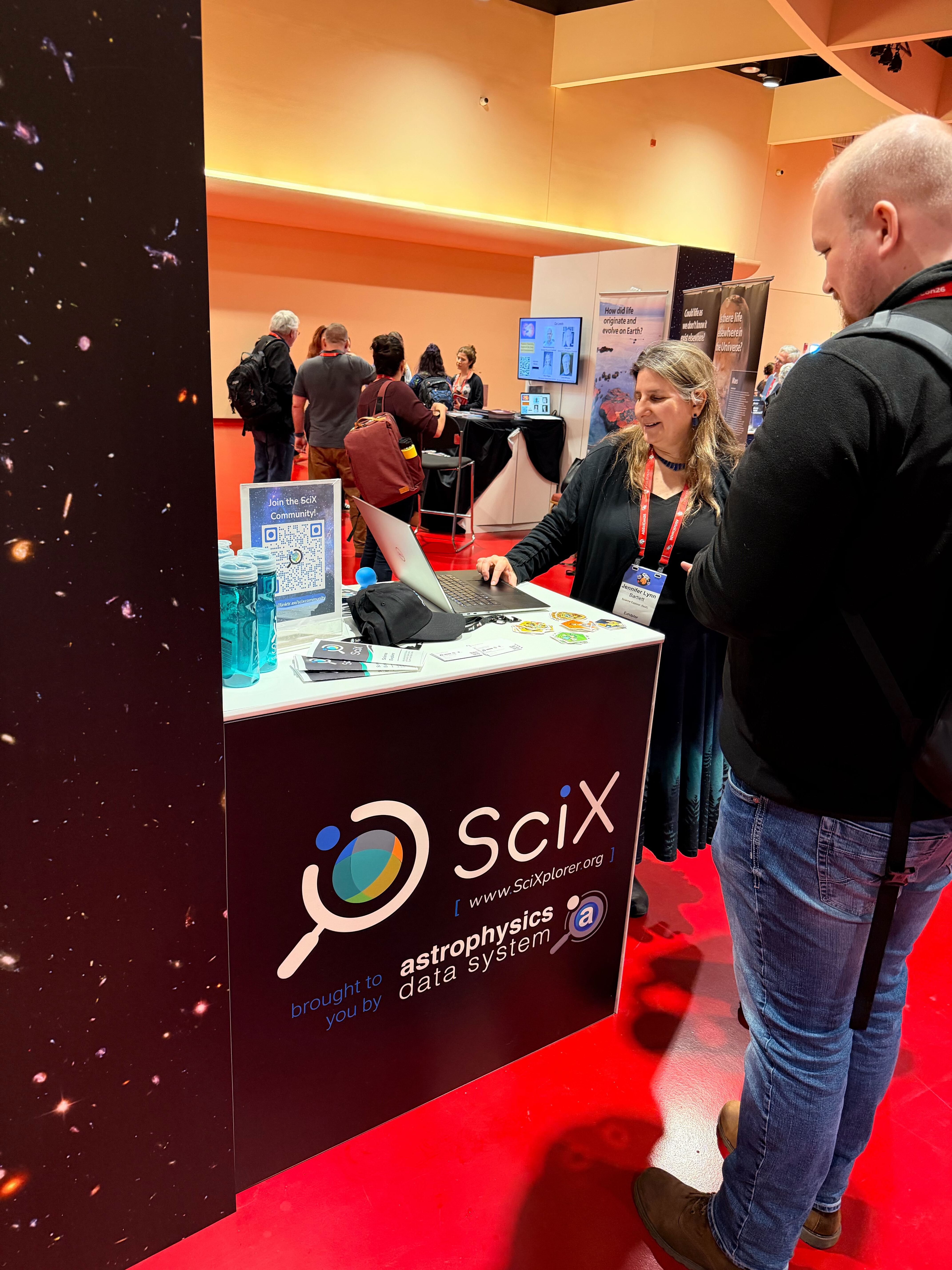 SciX team member pointing to a laptop screen demonstrating the SciX platform to attendees standing around the SciX conference booth